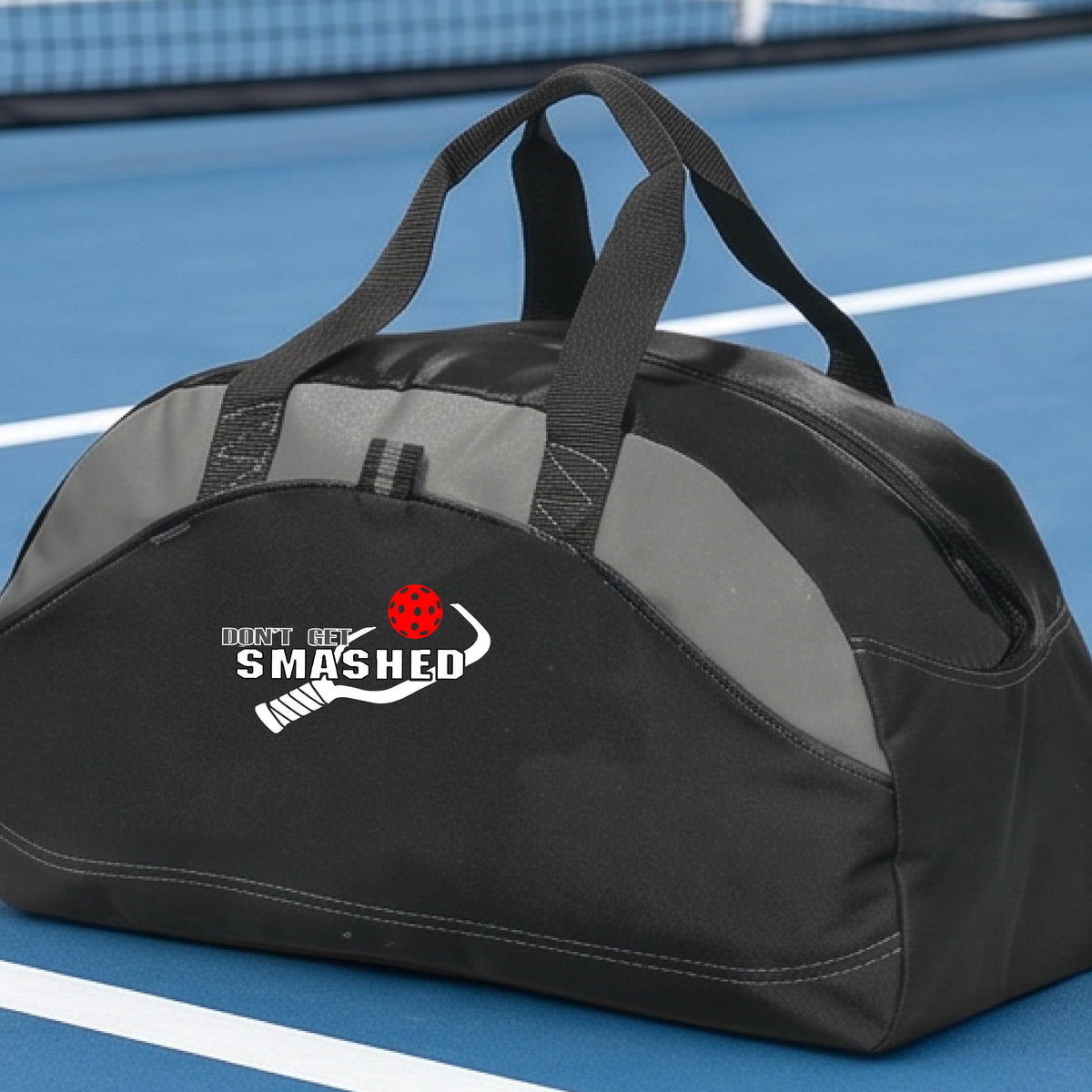 A close up of a black & grey duffel bag with white text "DON'T GET SMASHED" showing a white pickleball paddle and colorful picklball, sitting on a pickleball court. 