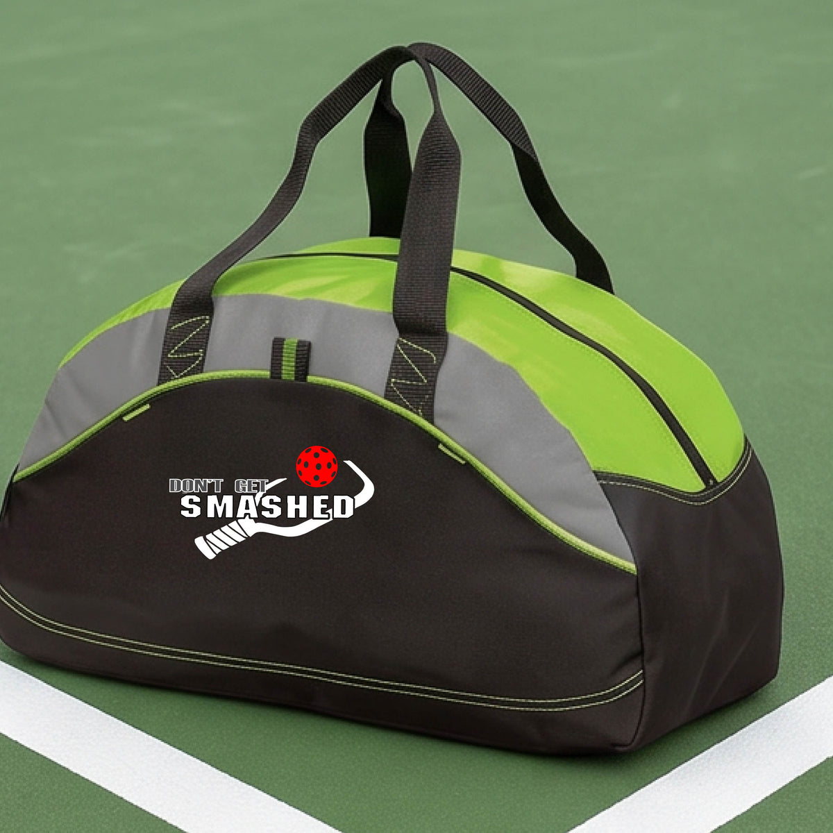 A close up of a lime green, black & grey duffel bag with white text "DON'T GET SMASHED" showing a white pickleball paddle and colorful picklball, sitting on a pickleball court. 
