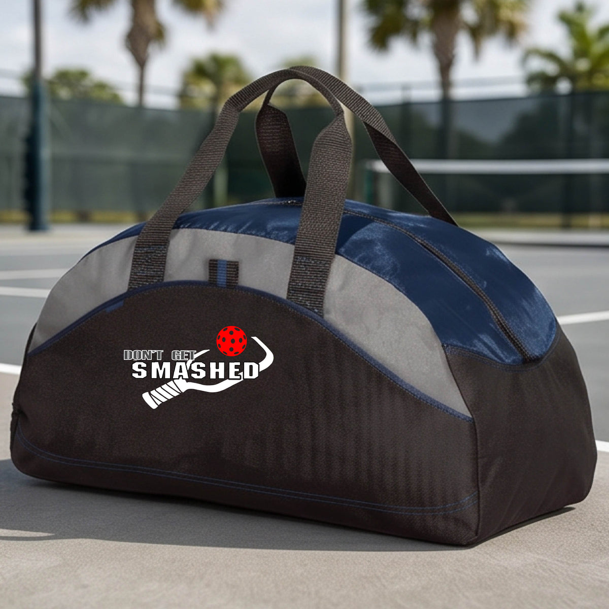 A close up of a navy blue, black & grey duffel bag with white text "DON'T GET SMASHED" showing a white pickleball paddle and colorful picklball, sitting on a pickleball court. 