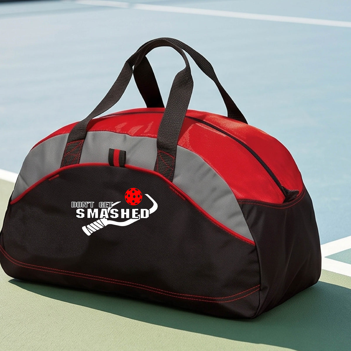 A close up of a red, black & grey duffel bag with white text "DON'T GET SMASHED" showing a white pickleball paddle and colorful picklball, sitting on a pickleball court. 