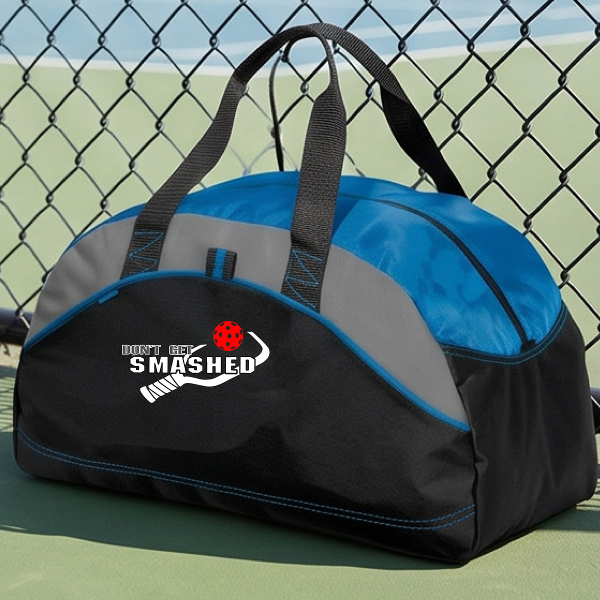 A close up of a royal blue, black & grey duffel bag with white text "DON'T GET SMASHED" showing a white pickleball paddle and colorful picklball, sitting on a pickleball court. 