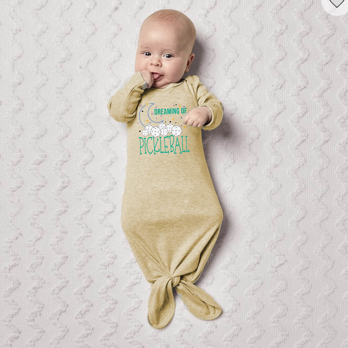 A baby wearing a beige long-sleeve sleep sack with a tie-front hem and 'DREAMING OF PICKLEBALL' text on the chest, lying on a textured white blanket.