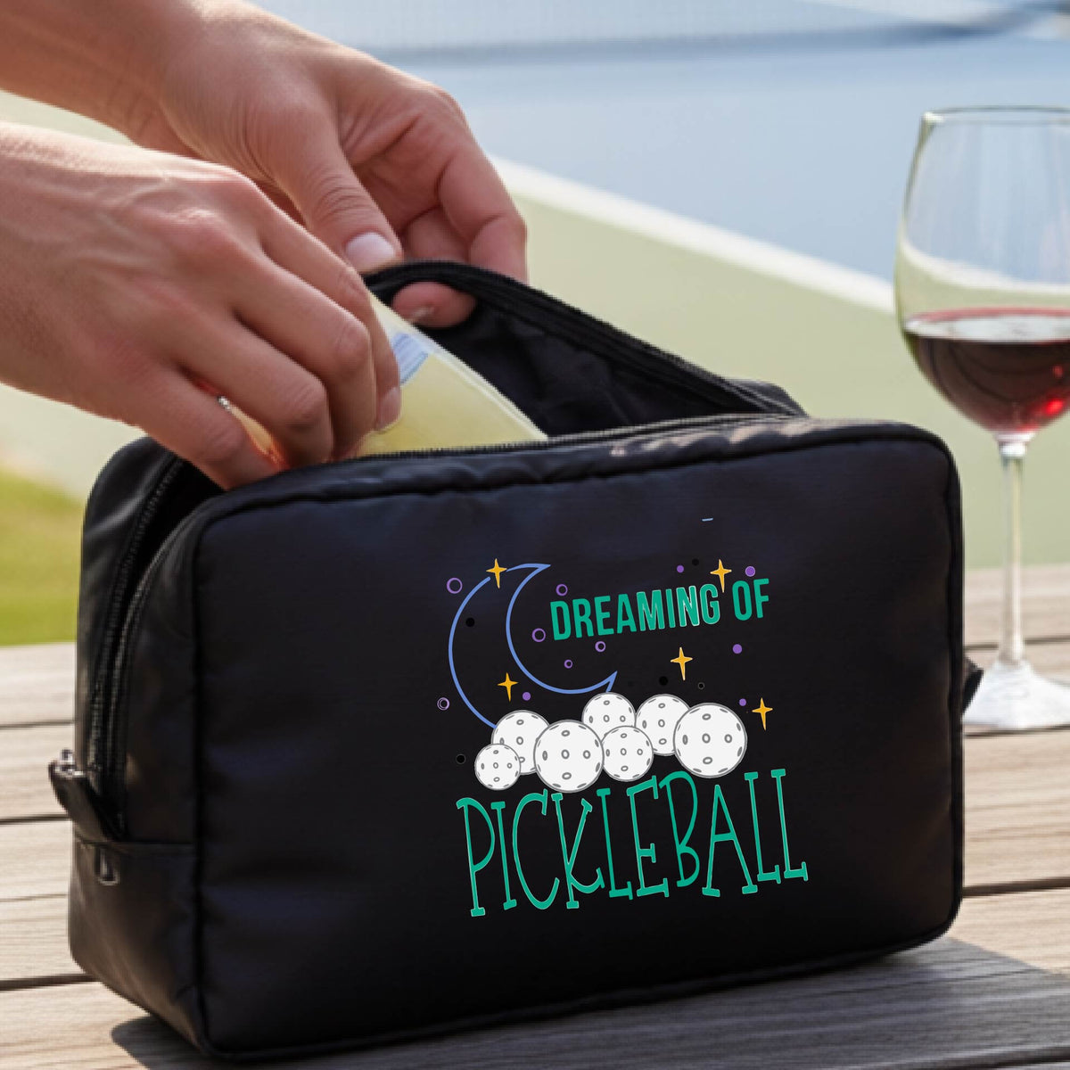 A black polyester cosmetic bag with a colorful 'Dreaming of Pickleball' graphic,with a pickleball court in the background