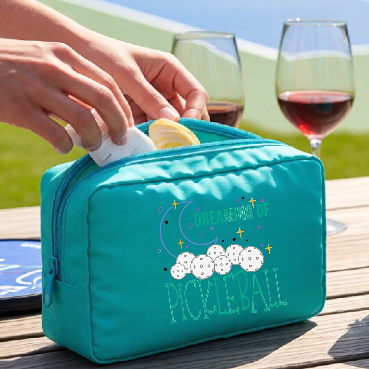 A tuquoise polyester cosmetic bag with a colorful 'Dreaming of Pickleball' graphic,with a pickleball court in the background