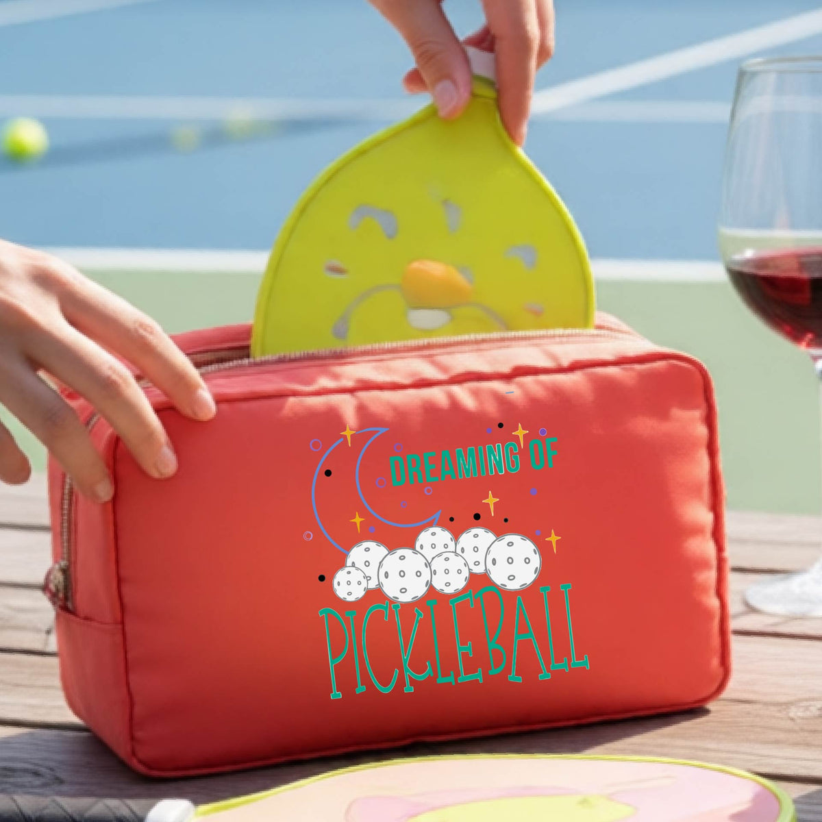 A coral colored polyester cosmetic bag with a colorful 'Dreaming of Pickleball' graphic,with a pickleball court in the background