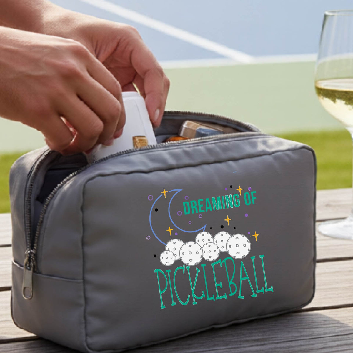 A gray polyester cosmetic bag with a colorful 'Dreaming of Pickleball' graphic,with a pickleball court in the background