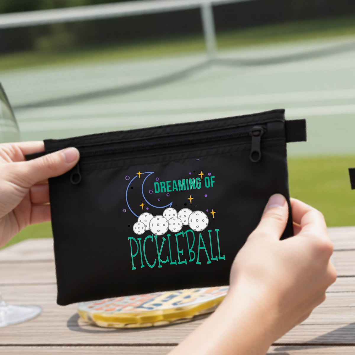 A black zippered pickleball pouch with a printed graphic of pickleballs, a crescent moon, and the words 'DREAMING OF PICKLEBALL.
