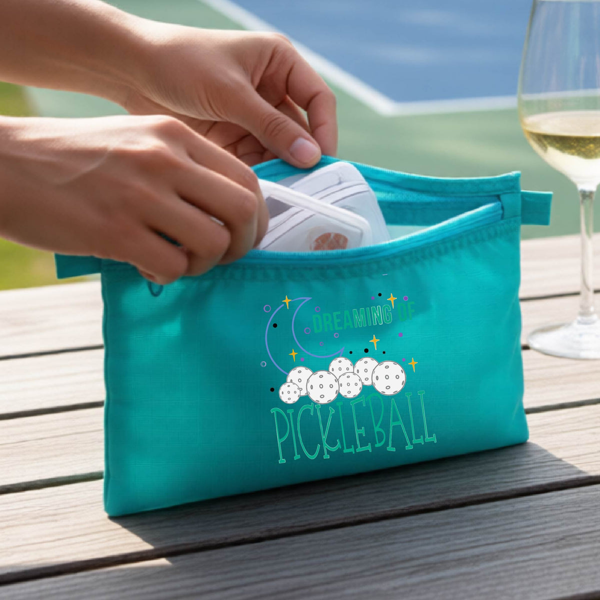 A turquoise zippered pickleball pouch with a printed graphic of pickleballs, a crescent moon, and the words 'DREAMING OF PICKLEBALL.