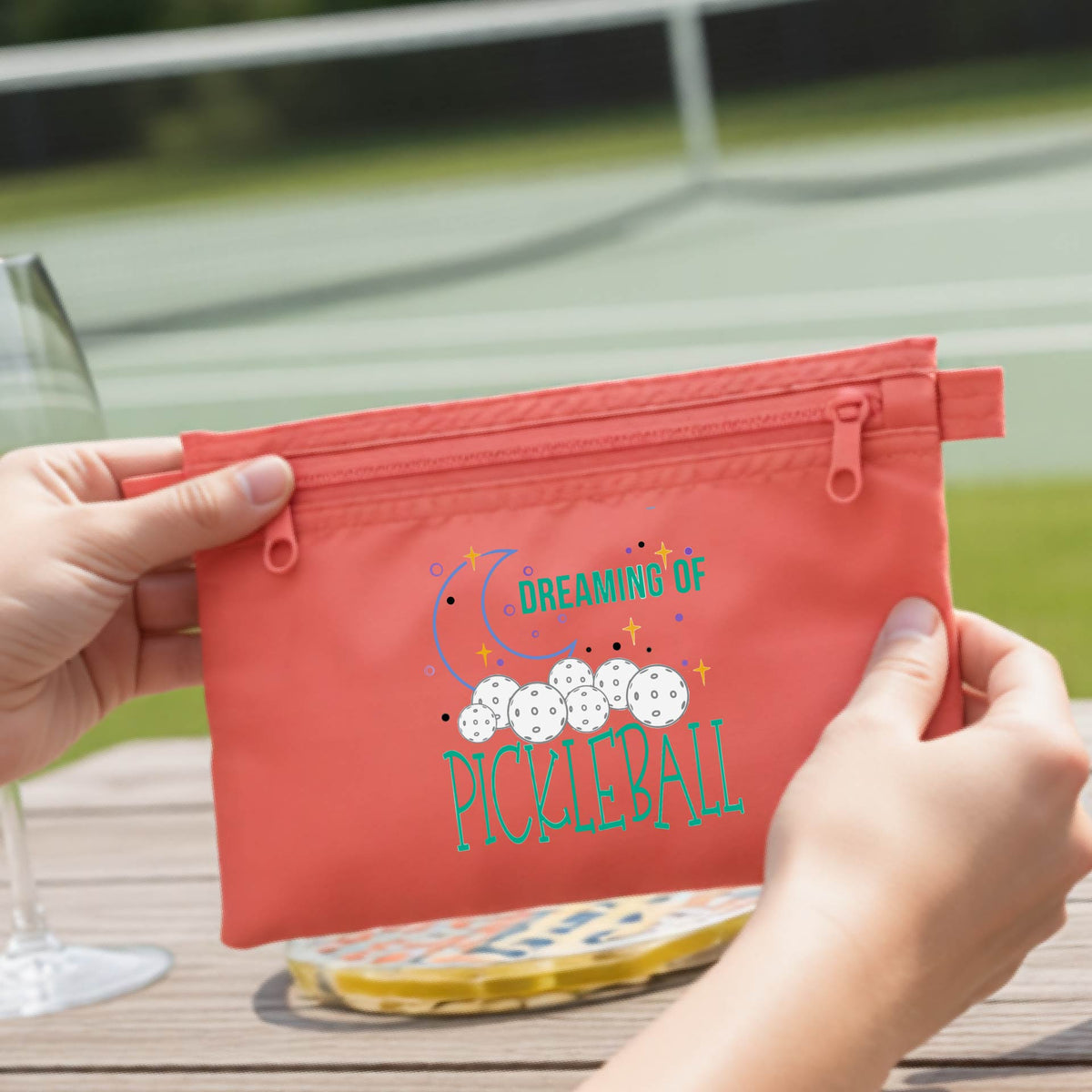 A coral-colored zippered pickleball pouch with a printed graphic of pickleballs, a crescent moon, and the words 'DREAMING OF PICKLEBALL.