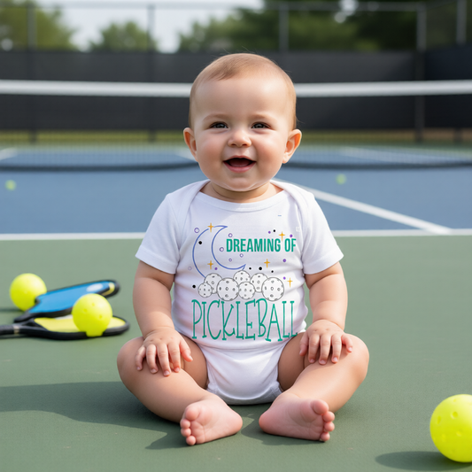 A baby sitting on a pickleball court wearing a white bodysuit with a colorful graphic that reads 'DREAMING OF PICKLEBALL'.
