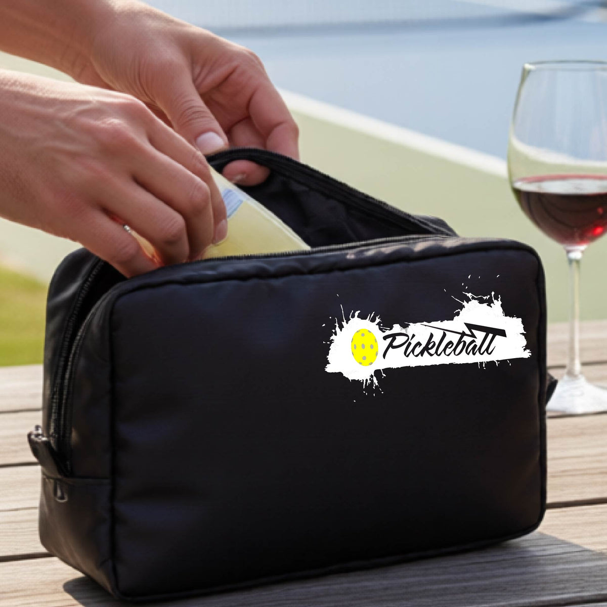 A black zippered pickleball-themed cosmetic bag with a white pickleball splaltter graphic and pickleball icon. 