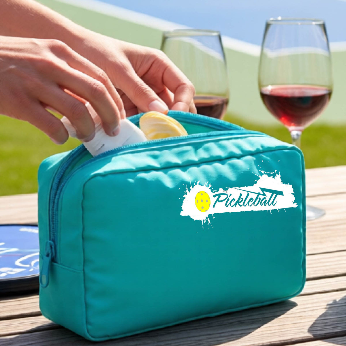 A turquoise zippered pickleball-themed cosmetic bag with a white pickleball splaltter graphic and pickleball icon. 