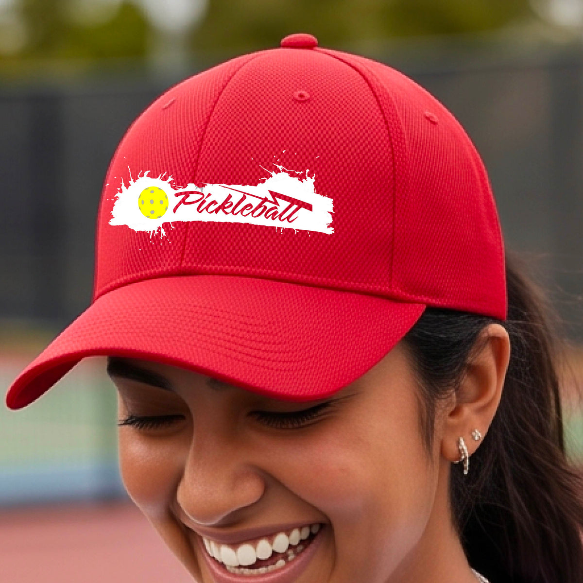 A close up of a red pickleball hat with a pickleball and white artistic splatter design with a pickleball court in the background. 