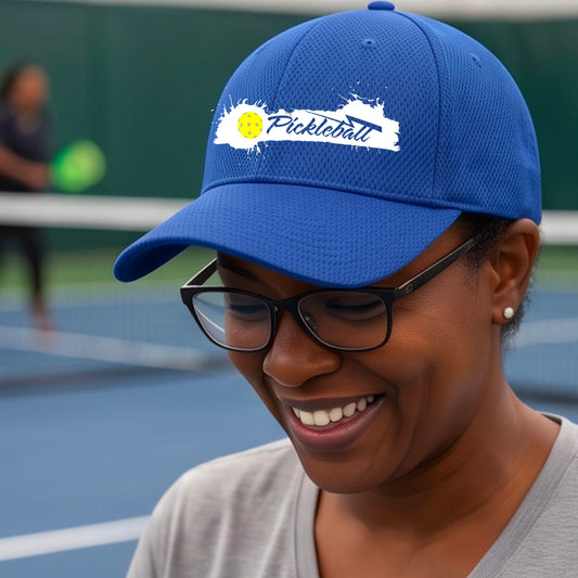 A close up of a royal blue pickleball hat with a pickleball and white artistic splatter design with a pickleball court in the background. 
