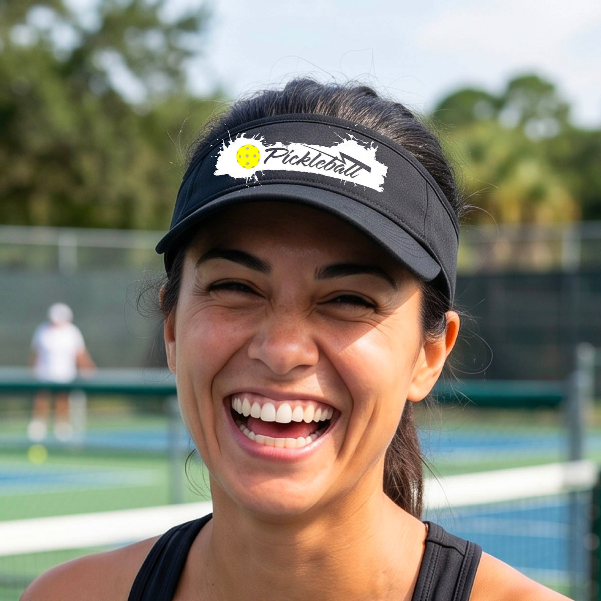 A smiling person wearing a black visor with a white paint-splatter logo that reads 'Pickleball' and a yellow pickleball graphic, photographed outdoors on a pickleball court.
