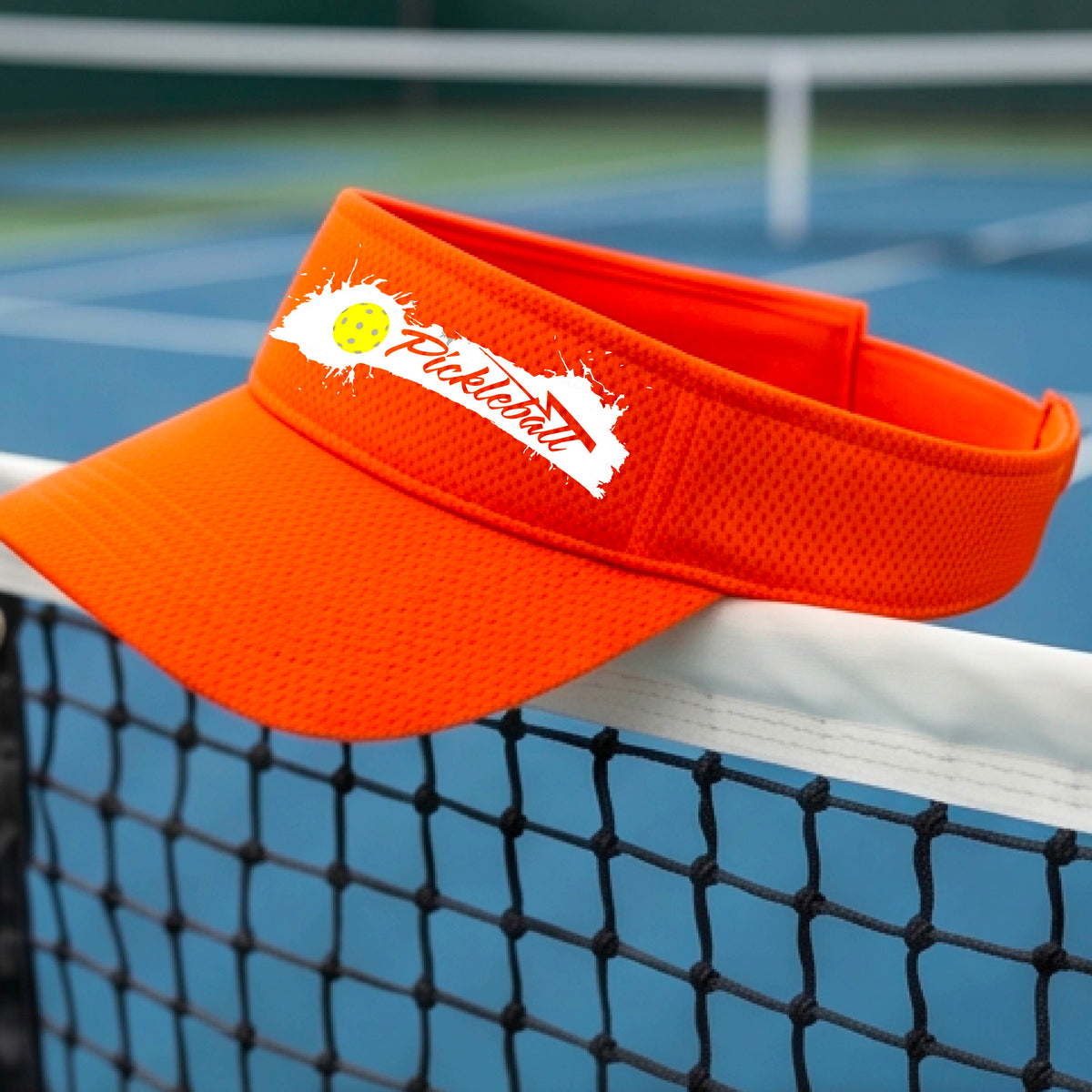 An orange visor with a white paint-splatter logo that reads 'Pickleball' and a yellow pickleball graphic, photographed outdoors on a pickleball court.