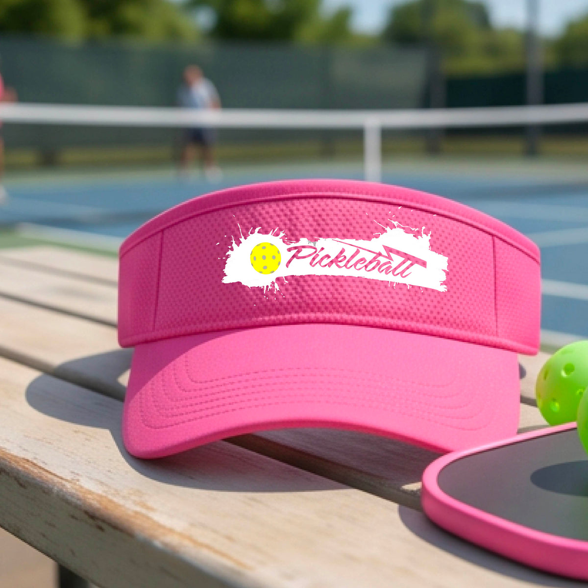 A pink visor with a white paint-splatter logo that reads 'Pickleball' and a yellow pickleball graphic, photographed outdoors on a pickleball court.