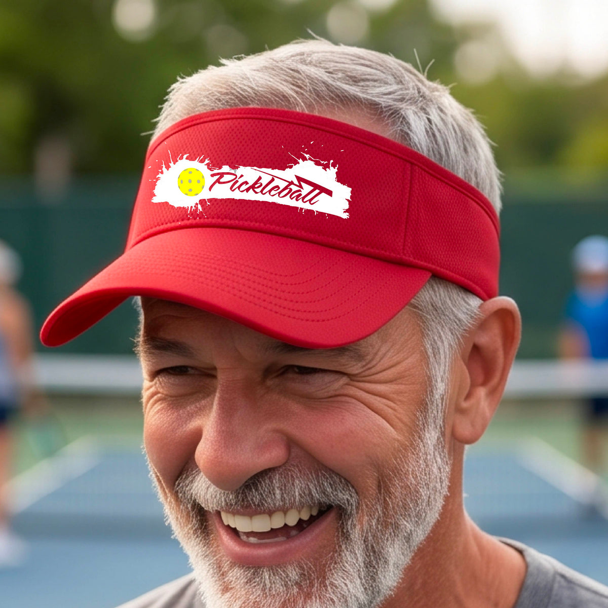 A smiling person wearing a red visor with a white paint-splatter logo that reads 'Pickleball' and a yellow pickleball graphic, photographed outdoors on a pickleball court.