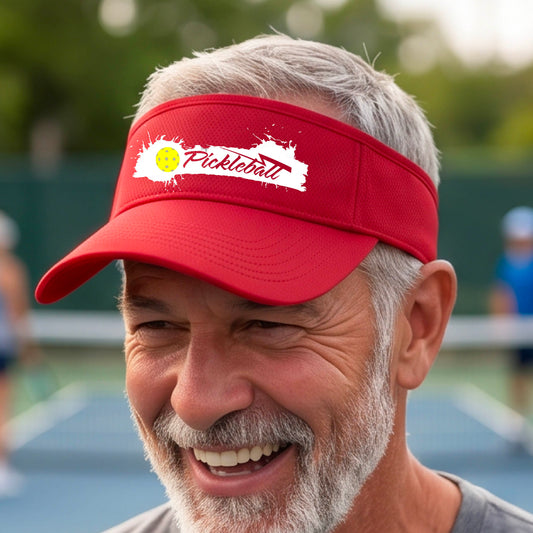 A smiling person wearing a red visor with a white paint-splatter logo that reads 'Pickleball' and a yellow pickleball graphic, photographed outdoors on a pickleball court.