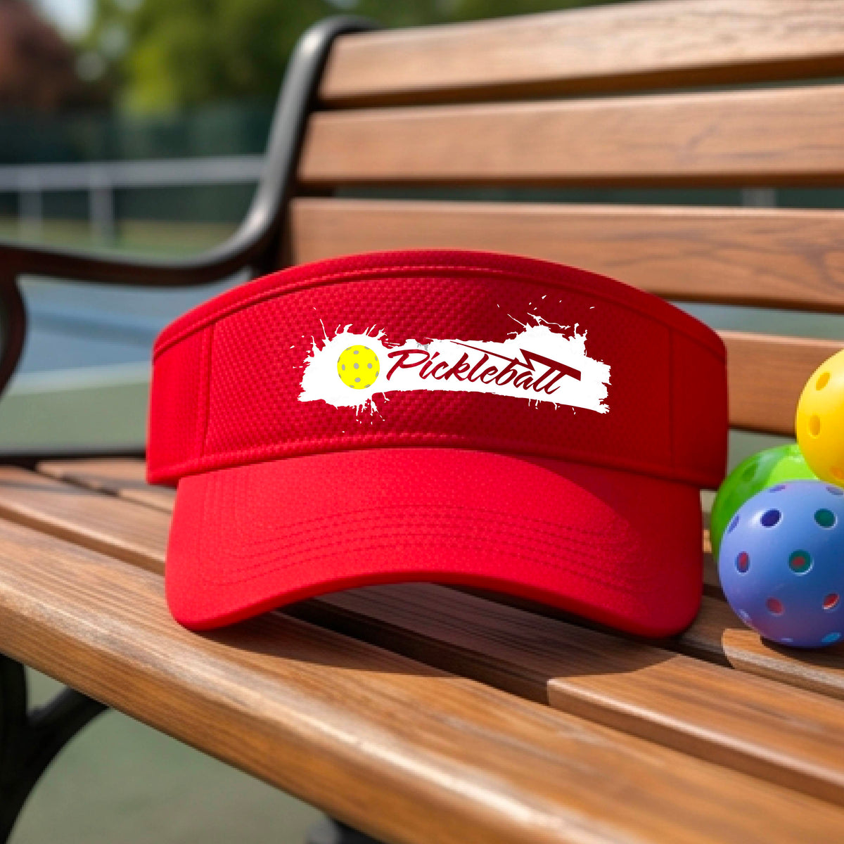 A red visor with a white paint-splatter logo that reads 'Pickleball' and a yellow pickleball graphic, photographed outdoors on a pickleball court.