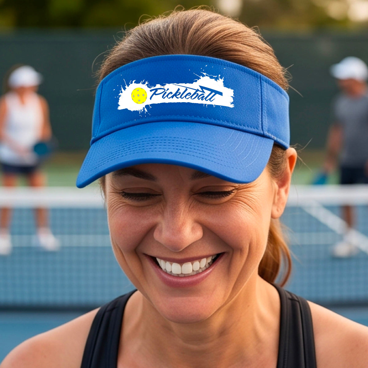 A smiling person wearing a royal blue visor with a white paint-splatter logo that reads 'Pickleball' and a yellow pickleball graphic, photographed outdoors on a pickleball court.