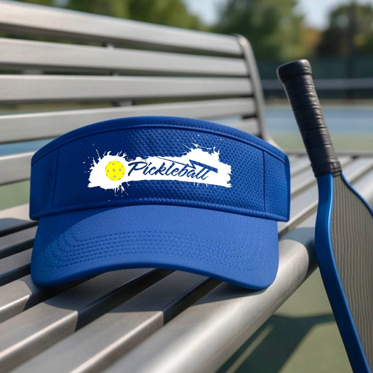 A royal blue visor with a white paint-splatter logo that reads 'Pickleball' and a yellow pickleball graphic, photographed outdoors on a pickleball court.