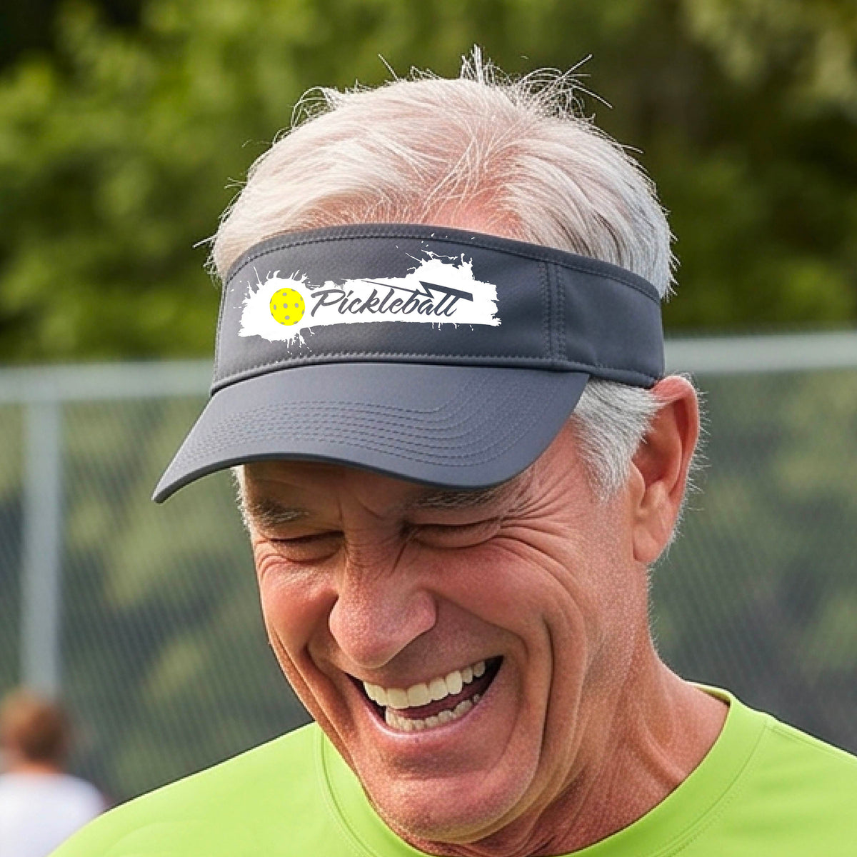 A smiling person wearing a gray visor with a white paint-splatter logo that reads 'Pickleball' and a yellow pickleball graphic, photographed outdoors on a pickleball court.