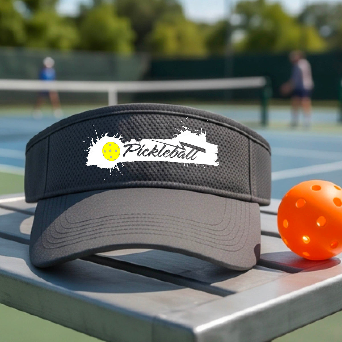 A gray visor with a white paint-splatter logo that reads 'Pickleball' and a yellow pickleball graphic, photographed outdoors on a pickleball court.