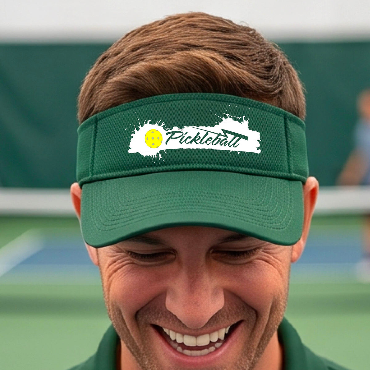 A smiling person wearing a green visor with a white paint-splatter logo that reads 'Pickleball' and a yellow pickleball graphic, photographed outdoors on a pickleball court.