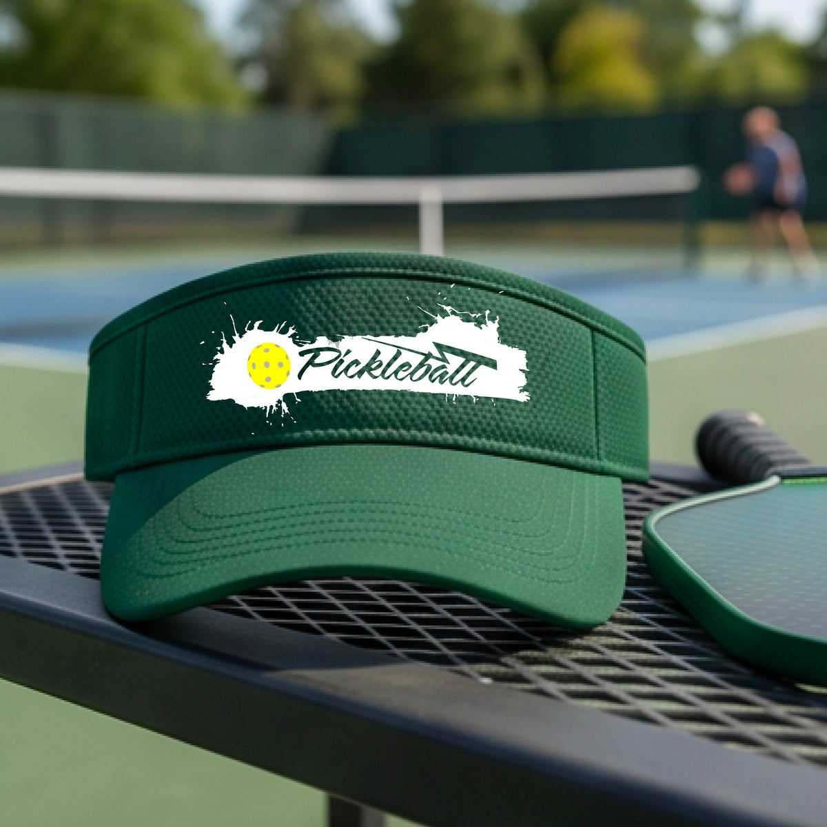 A green visor with a white paint-splatter logo that reads 'Pickleball' and a yellow pickleball graphic, photographed outdoors on a pickleball court.
