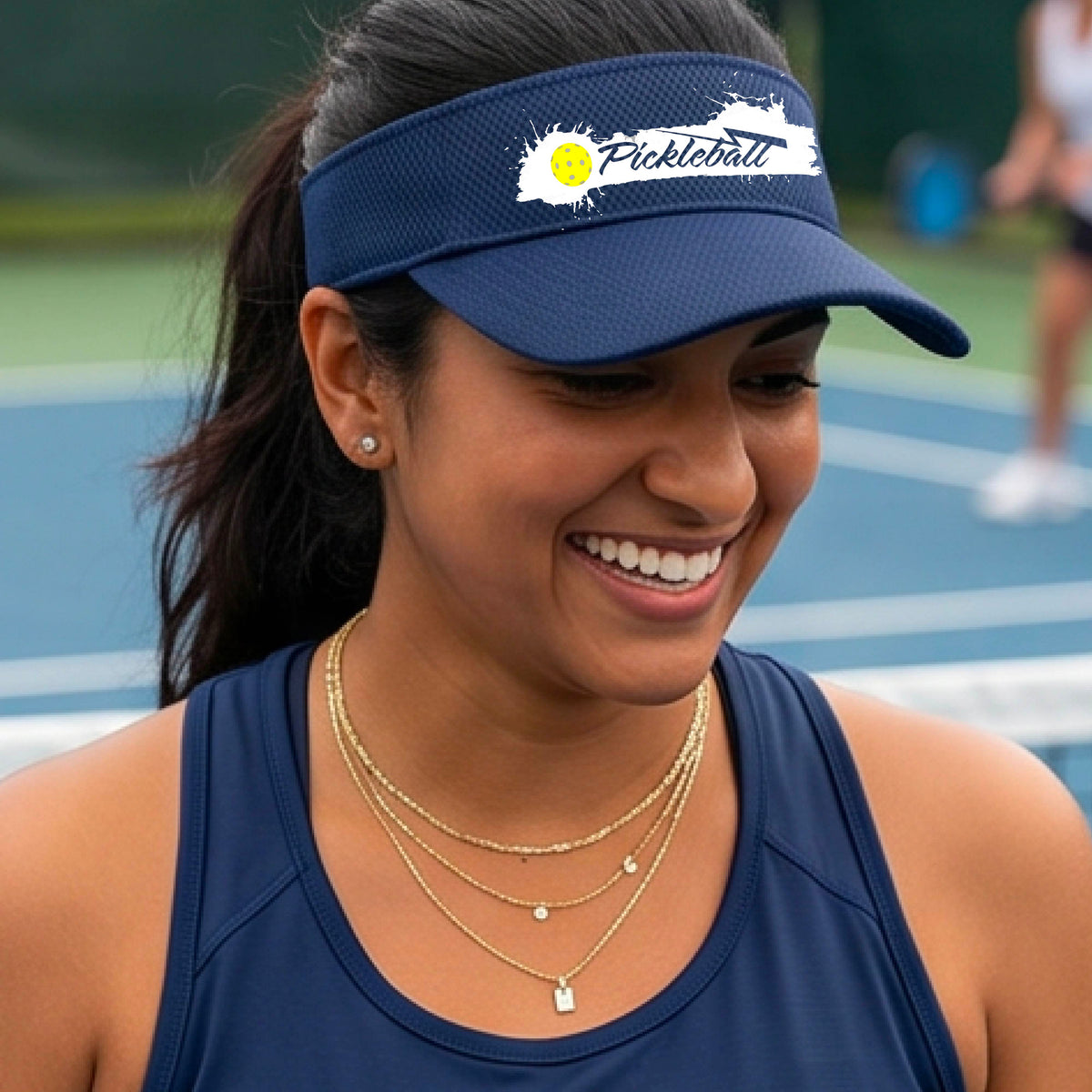 A smiling person wearing a navy blue visor with a white paint-splatter logo that reads 'Pickleball' and a yellow pickleball graphic, photographed outdoors on a pickleball court.