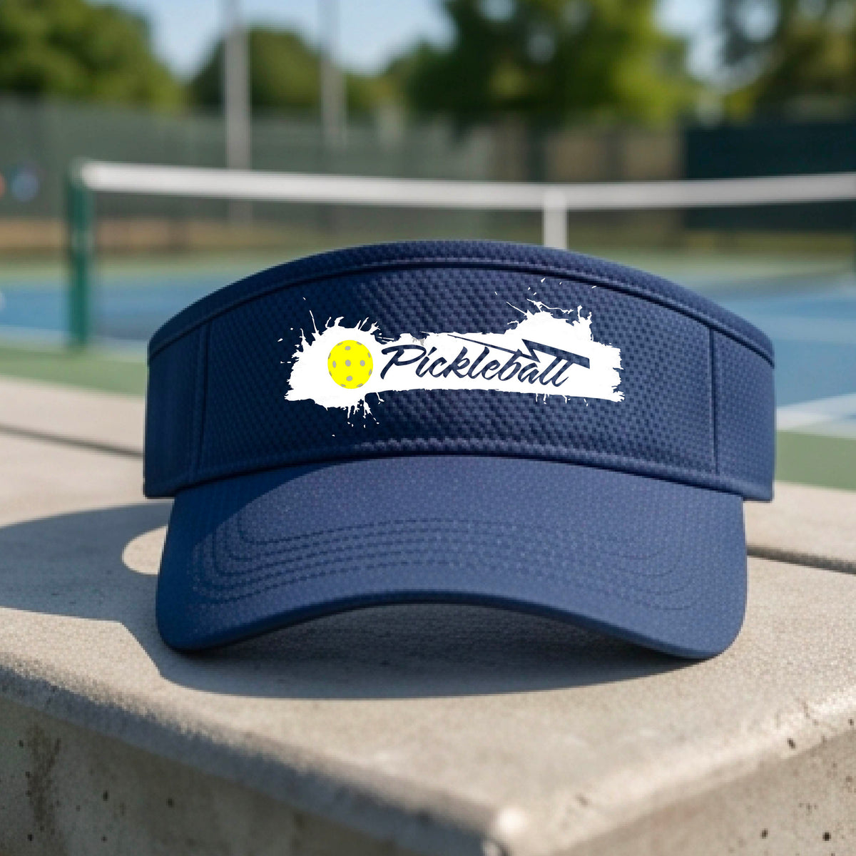 A navy blue visor with a white paint-splatter logo that reads 'Pickleball' and a yellow pickleball graphic, photographed outdoors on a pickleball court.