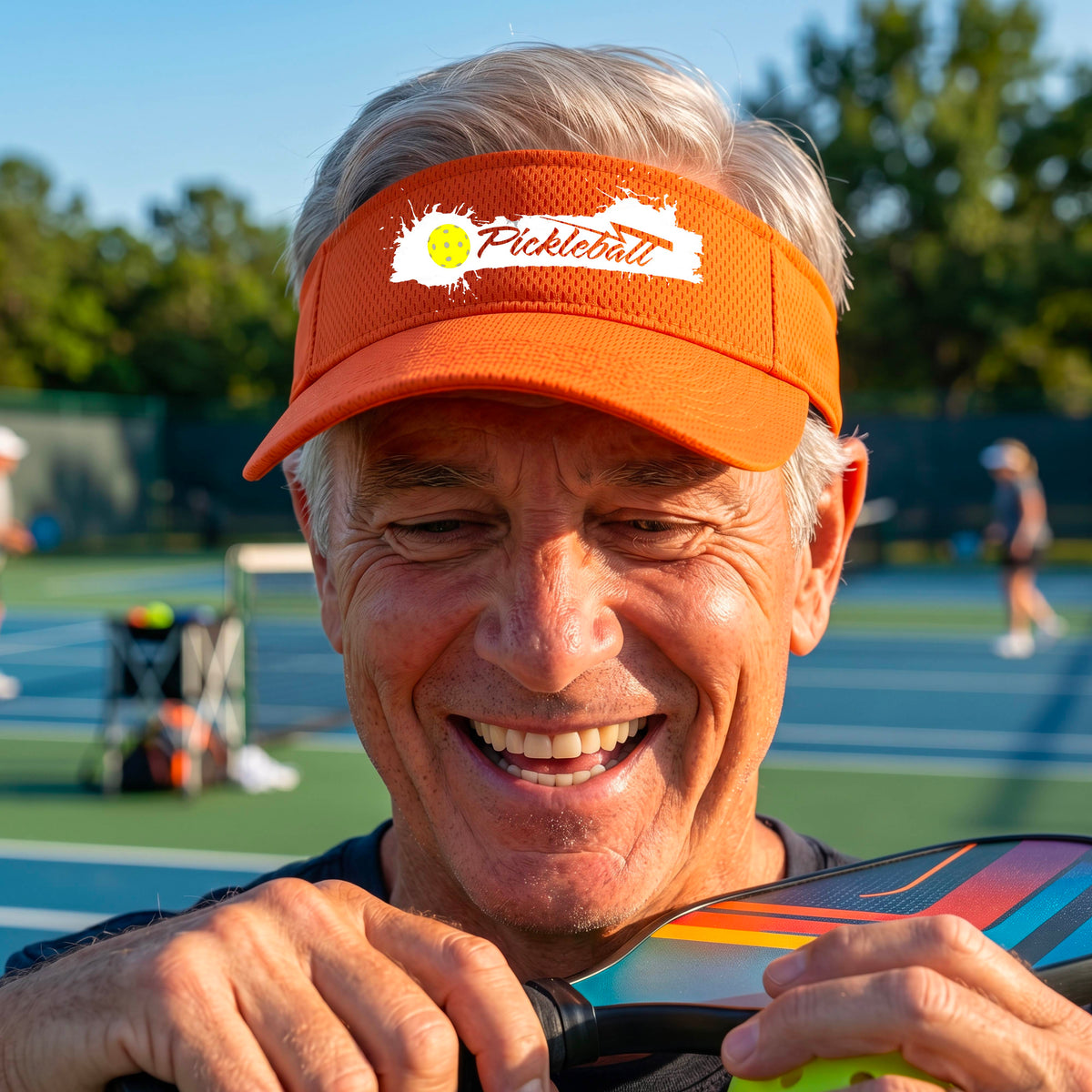 A smiling person wearing an orange visor with a white paint-splatter logo that reads 'Pickleball' and a yellow pickleball graphic, photographed outdoors on a pickleball court.