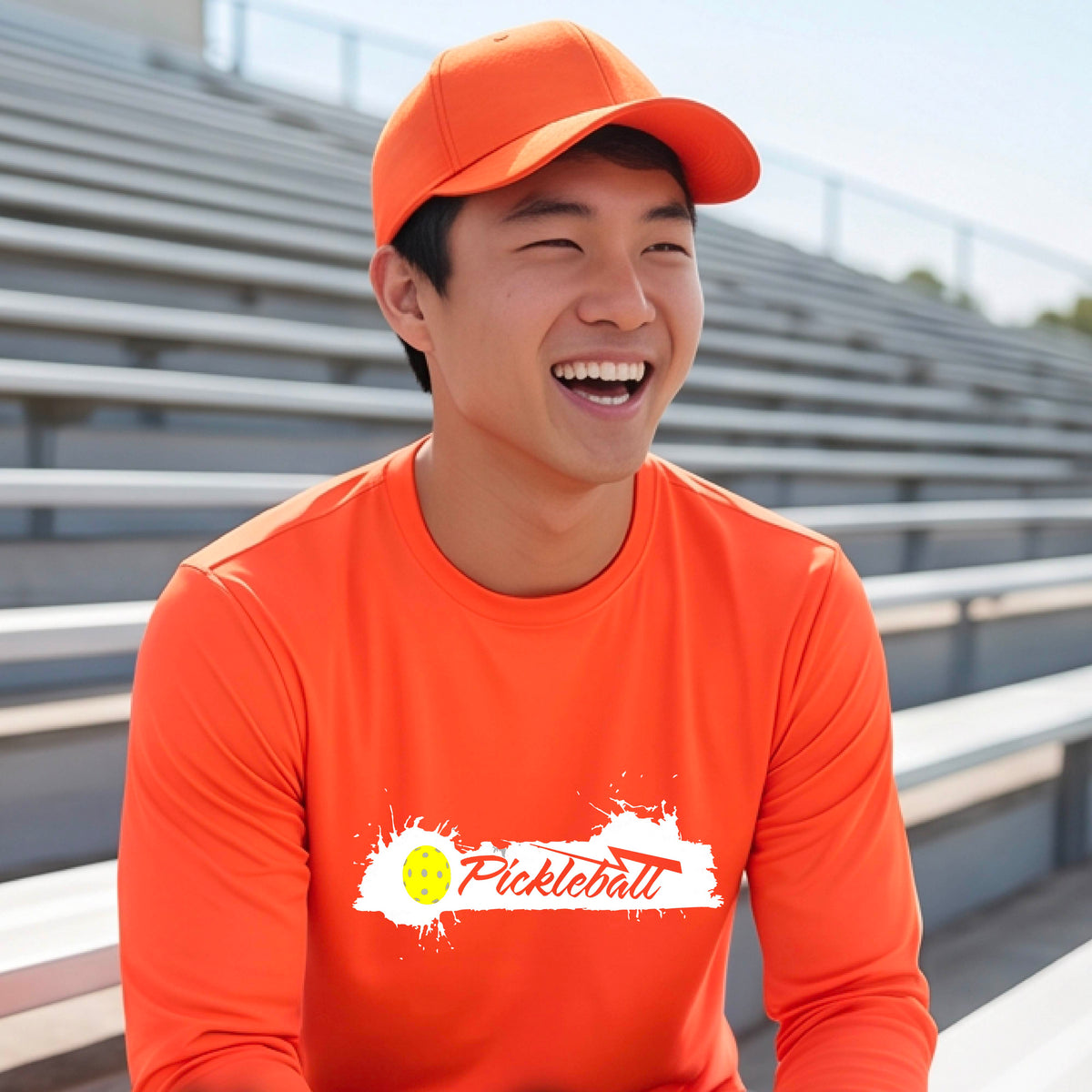 A smiling man wearing an orange long-sleeve t-shirt with a 'Pickleball' splatter graphic on the chest, standing on a pickleball court with players blurred in the background.