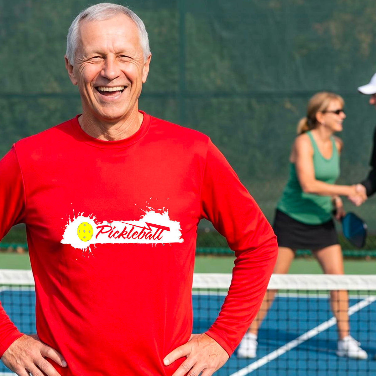 A smiling man wearing a red long-sleeve t-shirt with a 'Pickleball' splatter graphic on the chest, standing on a pickleball court with players blurred in the background.