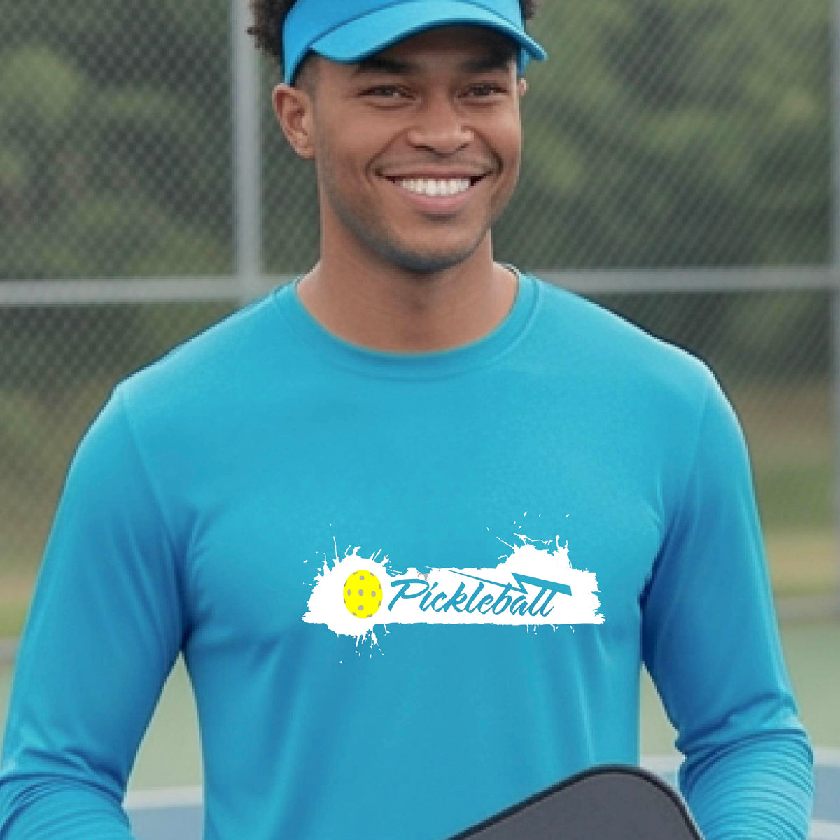 A smiling man wearing a bright blue long-sleeve t-shirt with a 'Pickleball' splatter graphic on the chest, standing on a pickleball court with players blurred in the background.