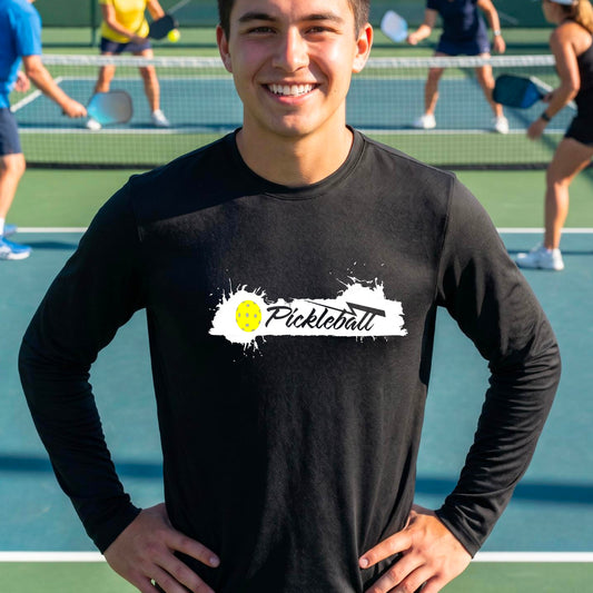 A smiling man wearing a black long-sleeve t-shirt with a 'Pickleball' splatter graphic on the chest, standing on a pickleball court with players blurred in the background.
