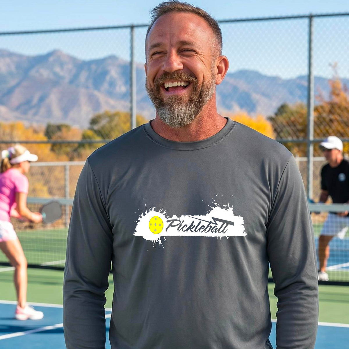 A smiling man wearing a gray long-sleeve t-shirt with a 'Pickleball' splatter graphic on the chest, standing on a pickleball court with players blurred in the background.