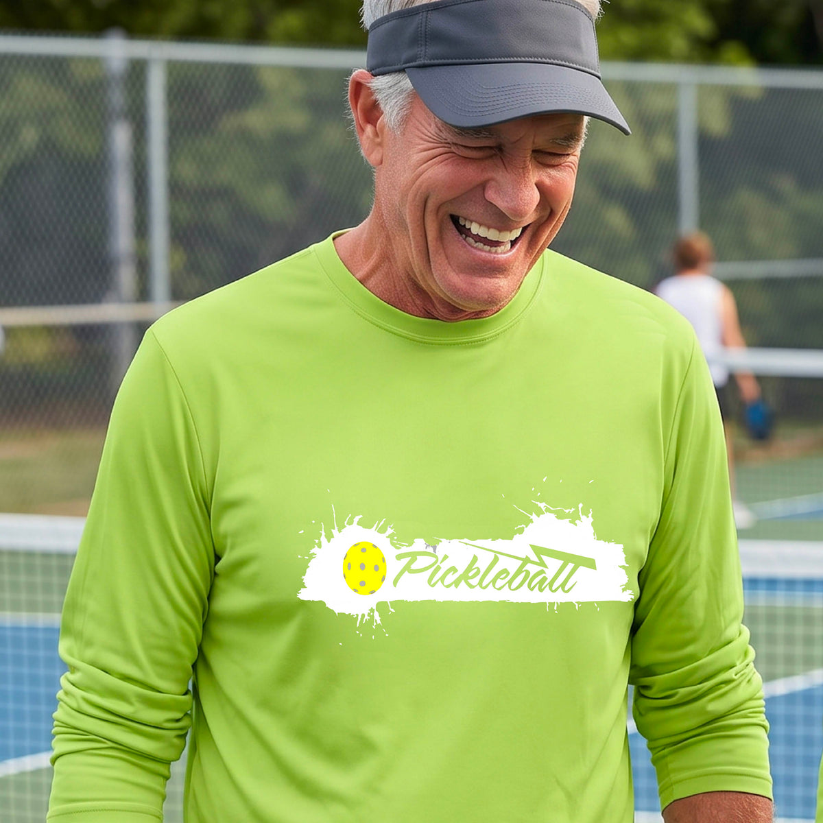 A smiling man wearing a lime green long-sleeve t-shirt with a 'Pickleball' splatter graphic on the chest, standing on a pickleball court with players blurred in the background.
