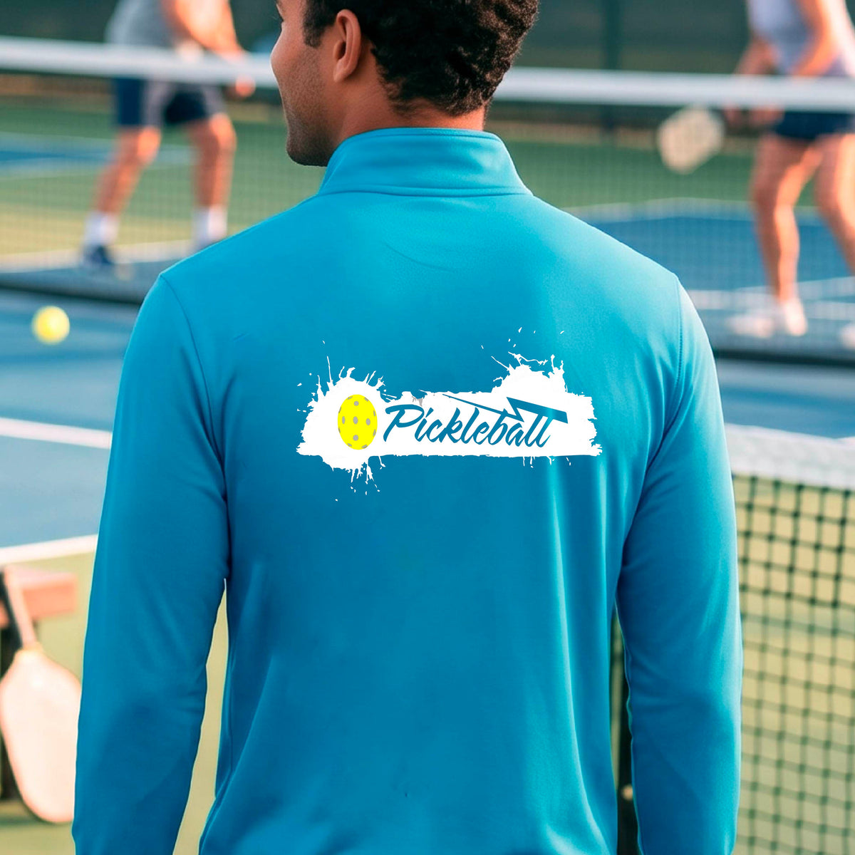 A man viewed from behind wearing a bright blue long-sleeve quarter-zip athletic shirt with a 'Pickleball' splatter  graphic standing on a pickleball court. 