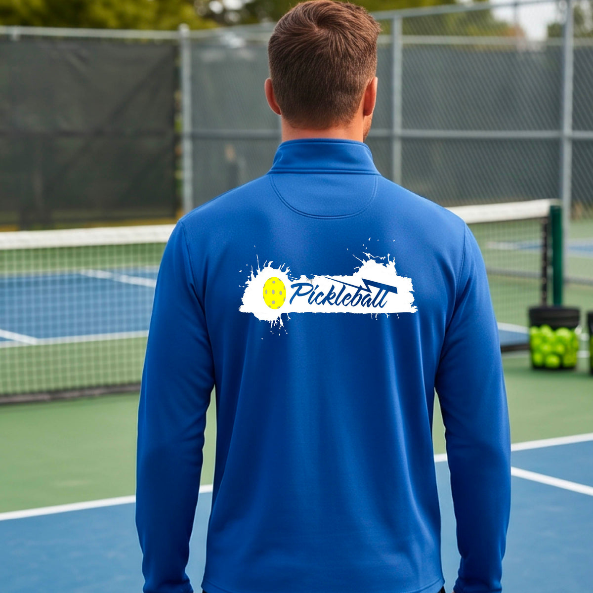 A man viewed from behind wearing a royal blue long-sleeve quarter-zip athletic shirt with a 'Pickleball' splatter  graphic standing on a pickleball court. 