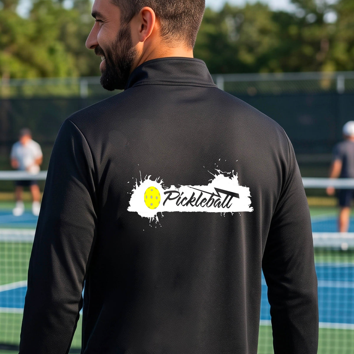 A man viewed from behind wearing a black long-sleeve quarter-zip athletic shirt with a 'Pickleball' splatter  graphic standing on a pickleball court. 