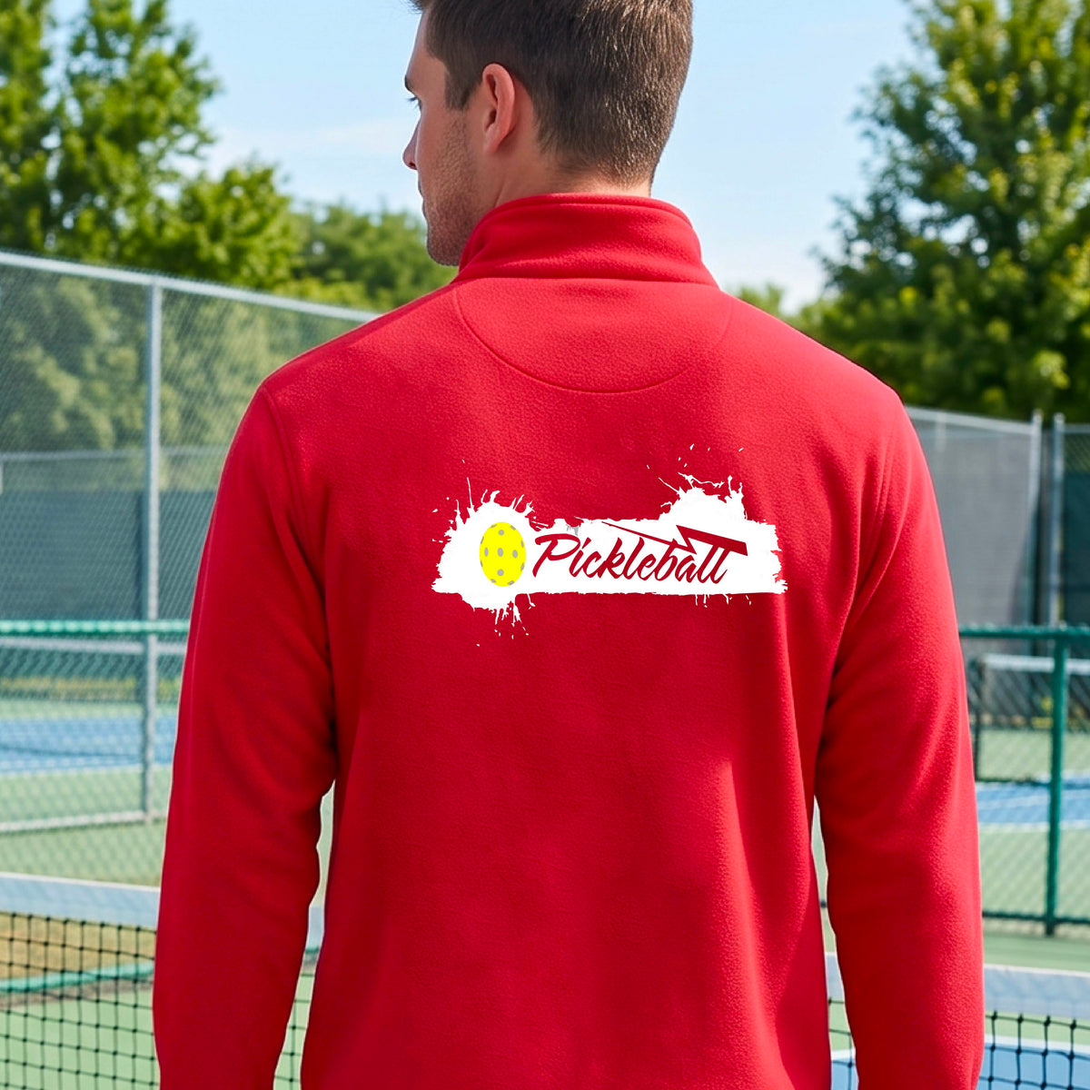 A man viewed from behind wearing a red long-sleeve quarter-zip athletic shirt with a 'Pickleball' splatter  graphic standing on a pickleball court. 