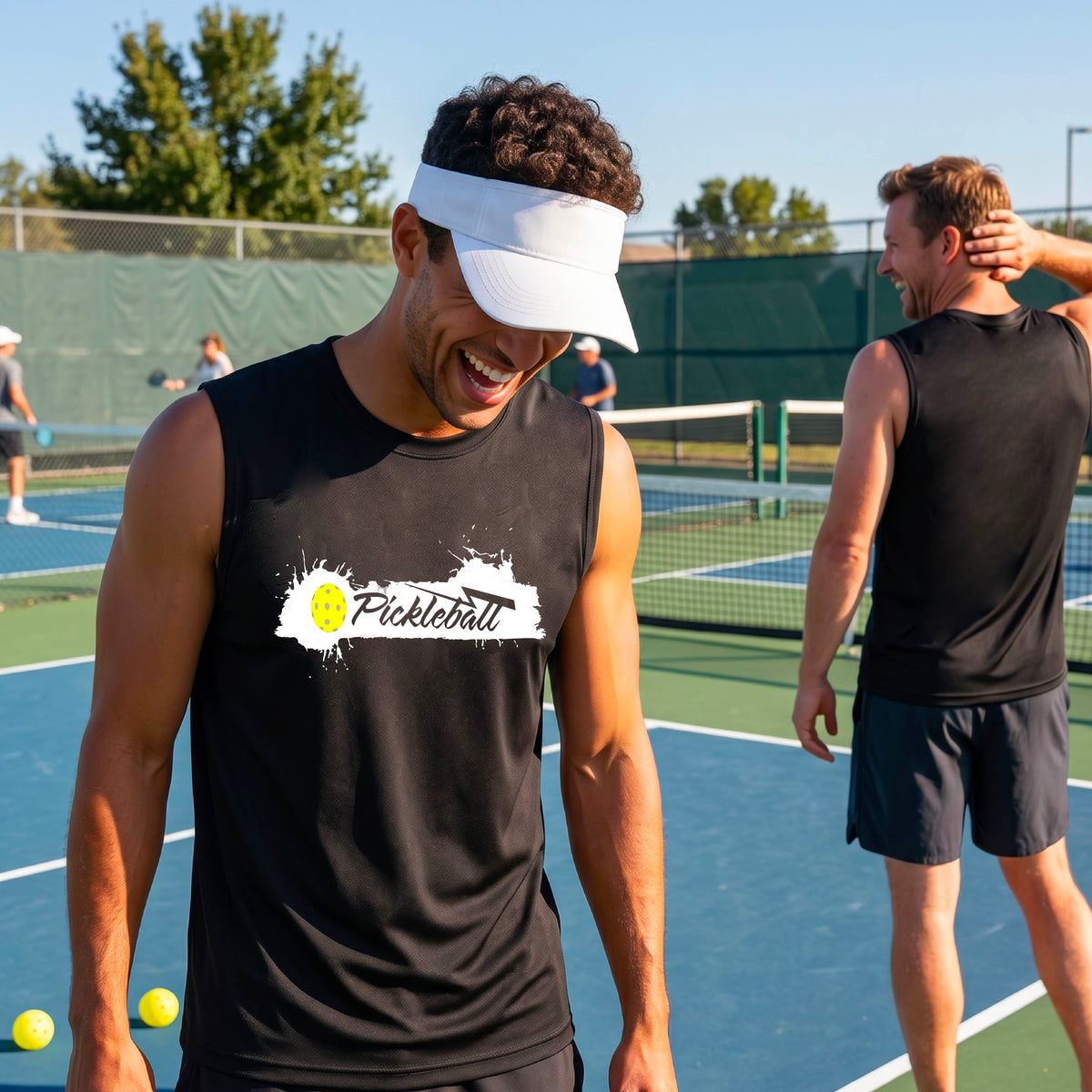 A smiling man wearing a black sleeveless athletic tank top with a 'Pickleball' splatter graphic stands on an outdoor pickleball court. 