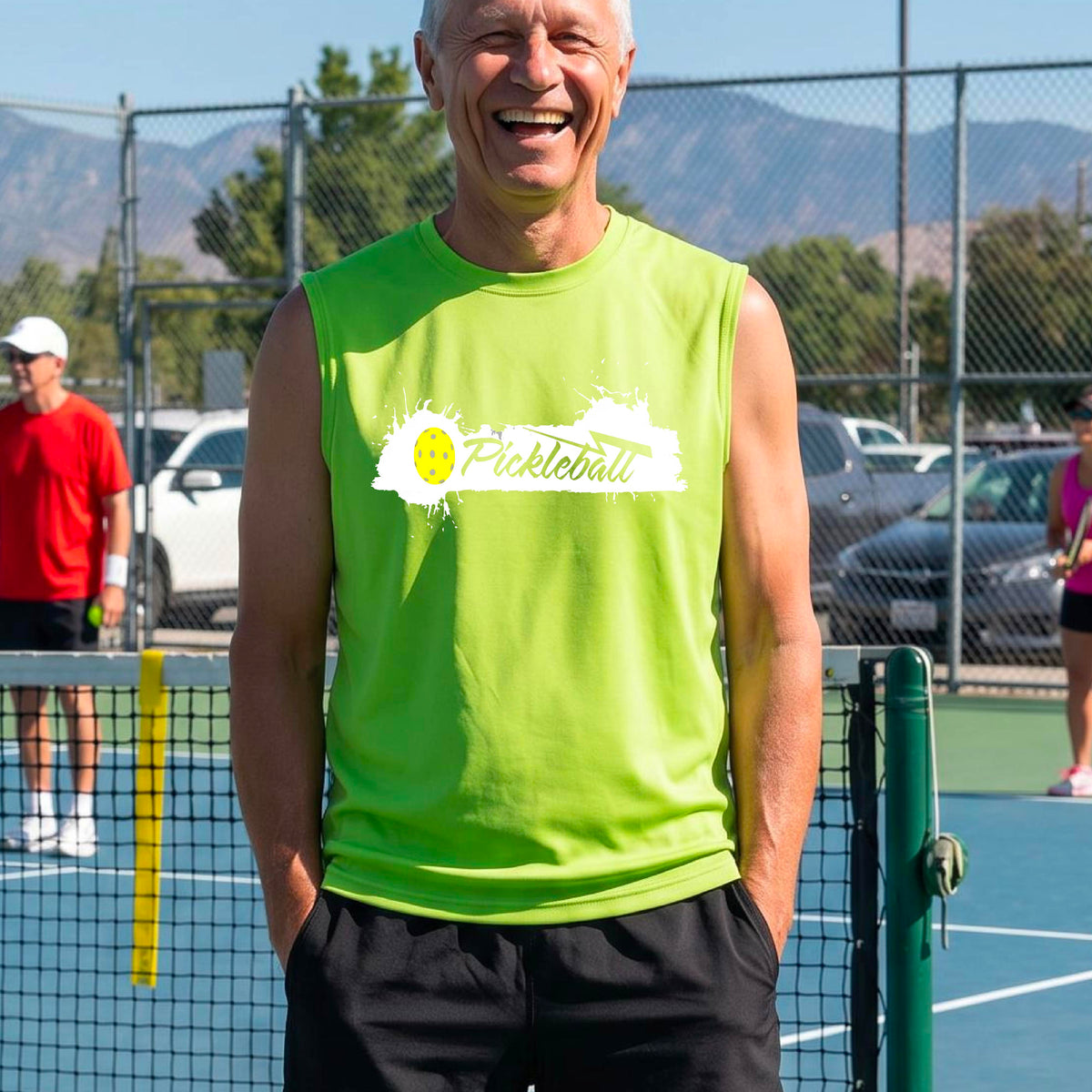 A smiling man wearing a lime green sleeveless athletic tank top with a 'Pickleball' splatter graphic stands on an outdoor pickleball court. 