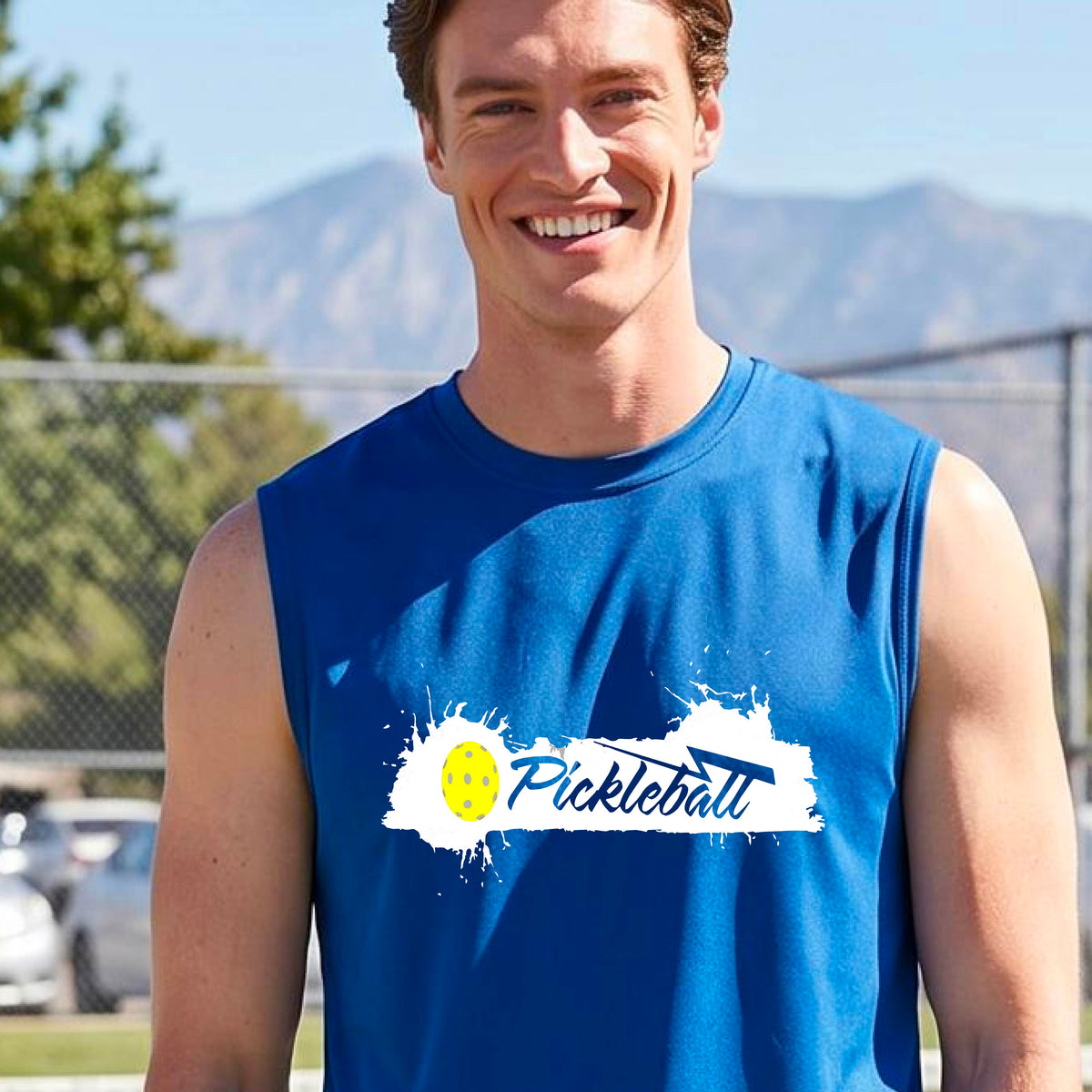A smiling man wearing a royal blue sleeveless athletic tank top with a 'Pickleball' splatter graphic stands on an outdoor pickleball court. 