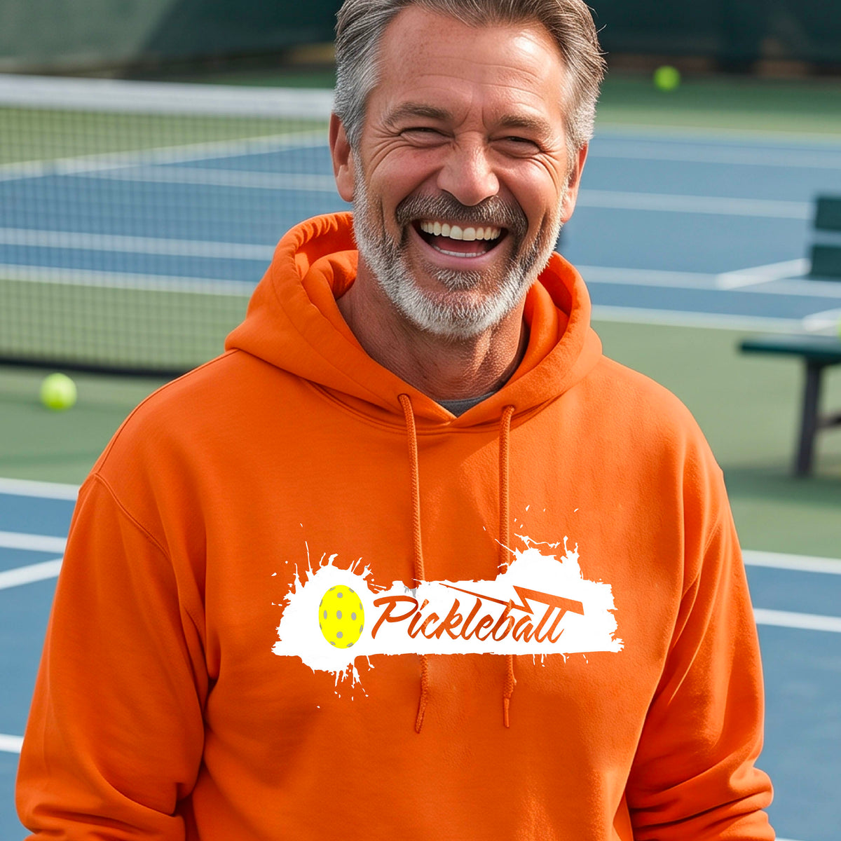 A smiling older man wearing a bright orange hoodie with a 'Pickleball' splatter graphic on the chest stands on an outdoor pickleball court. 