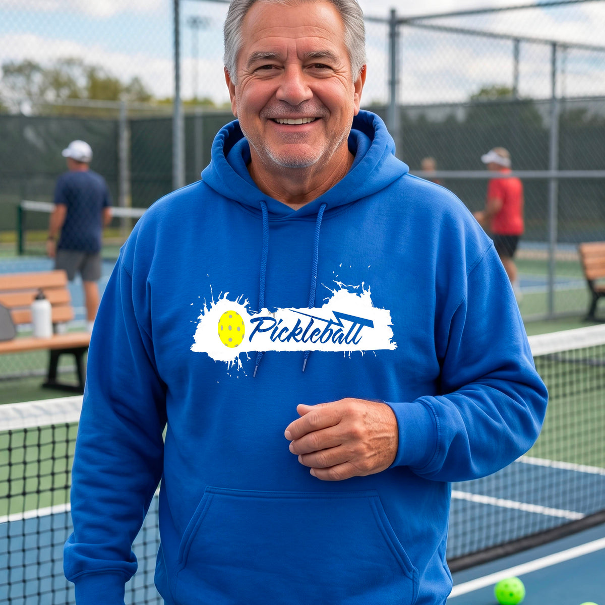 A smiling older man wearing a bright blue hoodie with a 'Pickleball' splatter graphic on the chest stands on an outdoor pickleball court. 
