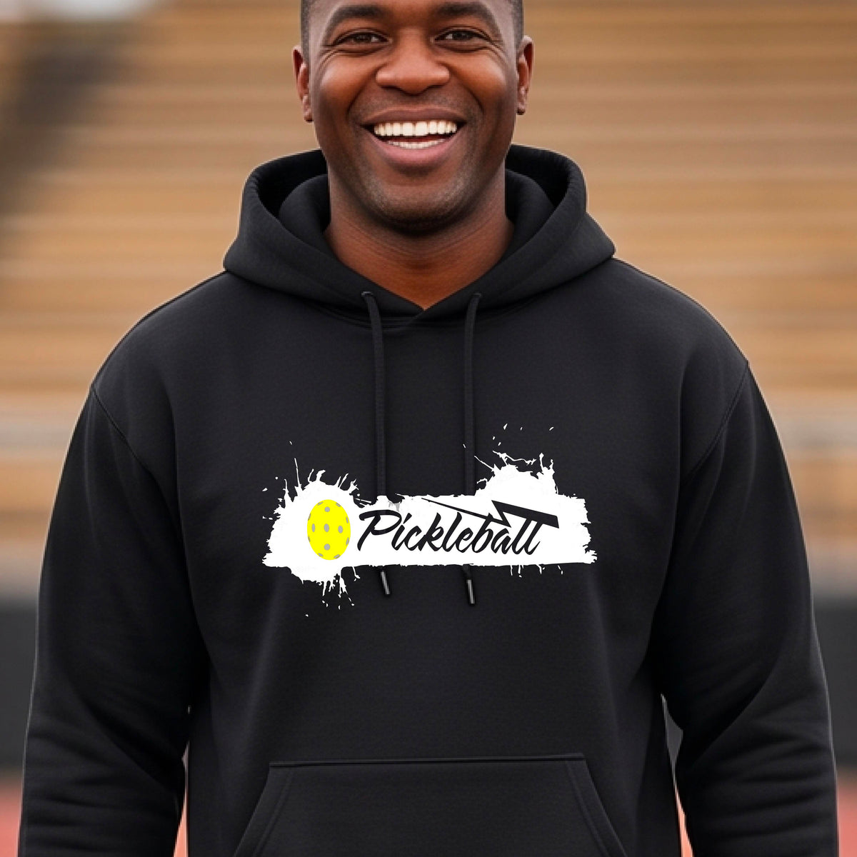 A smiling  man wearing a black hoodie with a 'Pickleball' splatter graphic on the chest stands on an outdoor pickleball court. 