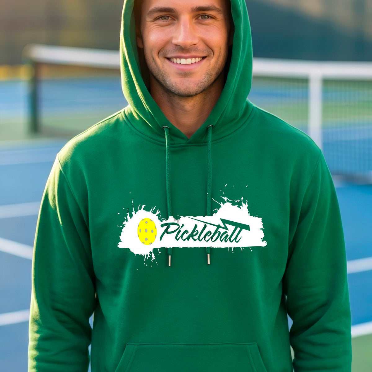 A smiling man wearing a green hoodie with a 'Pickleball' splatter graphic on the chest stands on an outdoor pickleball court. 